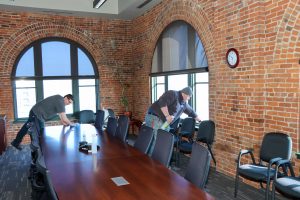 Two people cleaning a conference room with arched windows and exposed brick walls, featuring a long table and black chairs.