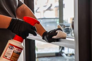 Person wearing black gloves cleaning a window with a spray bottle and cloth.