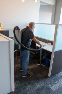 A janitor cleaning an office cubicle with a backpack vacuum, featuring modern office furniture and frosted glass dividers.