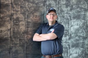 Man wearing glasses and a cap, standing with arms crossed in front of a dark textured wall.