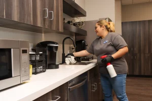 Person cleaning a kitchen countertop with a spray bottle and cloth, standing near a sink and coffee maker.